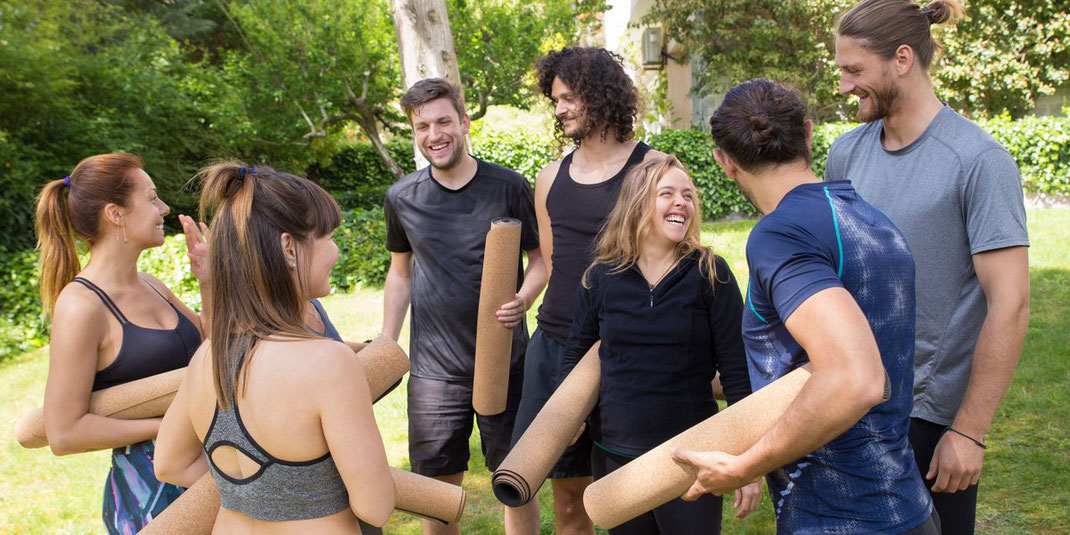 Eine fröhliche Gruppe von sechs Personen in Sportkleidung steht nach einem Yoga-Kurs in einem grünen Park. Alle halten zusammengerollte Yogamatten und unterhalten sich lachend. Die Gruppe besteht aus Männern und Frauen verschiedener Altersgruppen, alle i