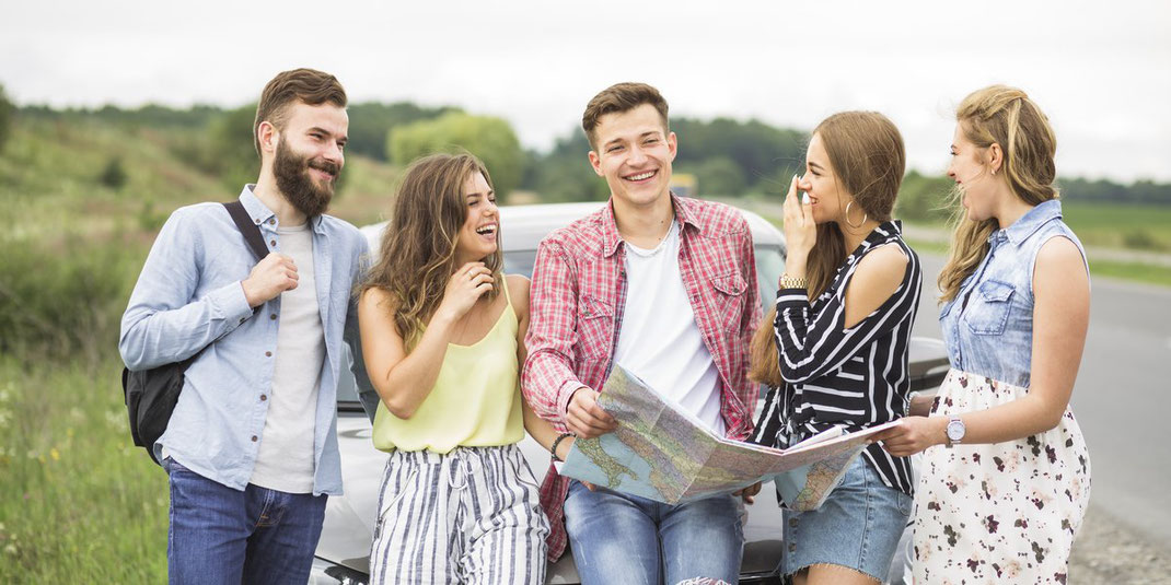 Gruppe junger Freunde steht lachend vor einem Auto, einer hält eine Landkarte in der Hand – Symbol für Abenteuer und Roadtrip.