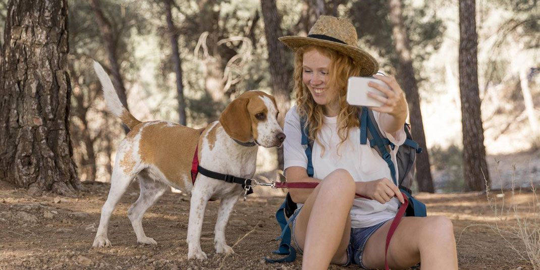 Junge Frau mit Strohhut sitzt im Wald und macht ein Selfie mit ihrem Hund an der Leine.