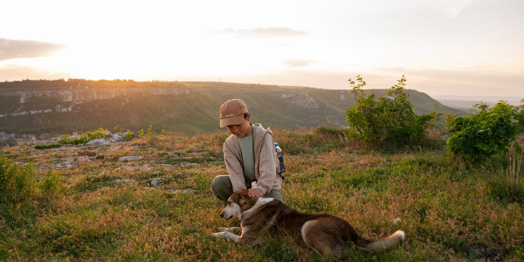 Kind in beiger Kappe hockt in der Natur bei Sonnenuntergang und streichelt einen liegenden Hund auf einer Wiese