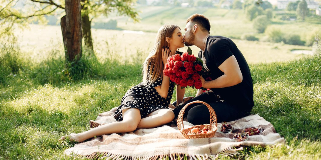 Verliebtes Paar küsst sich beim Picknick auf einer Decke, der Mann hält einen Strauß roter Rosen, umgeben von Obst und Natur.