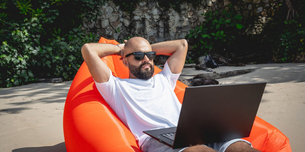 Mann mit Sonnenbrille arbeitet entspannt am Laptop auf einem aufblasbaren Sofa am Strand.