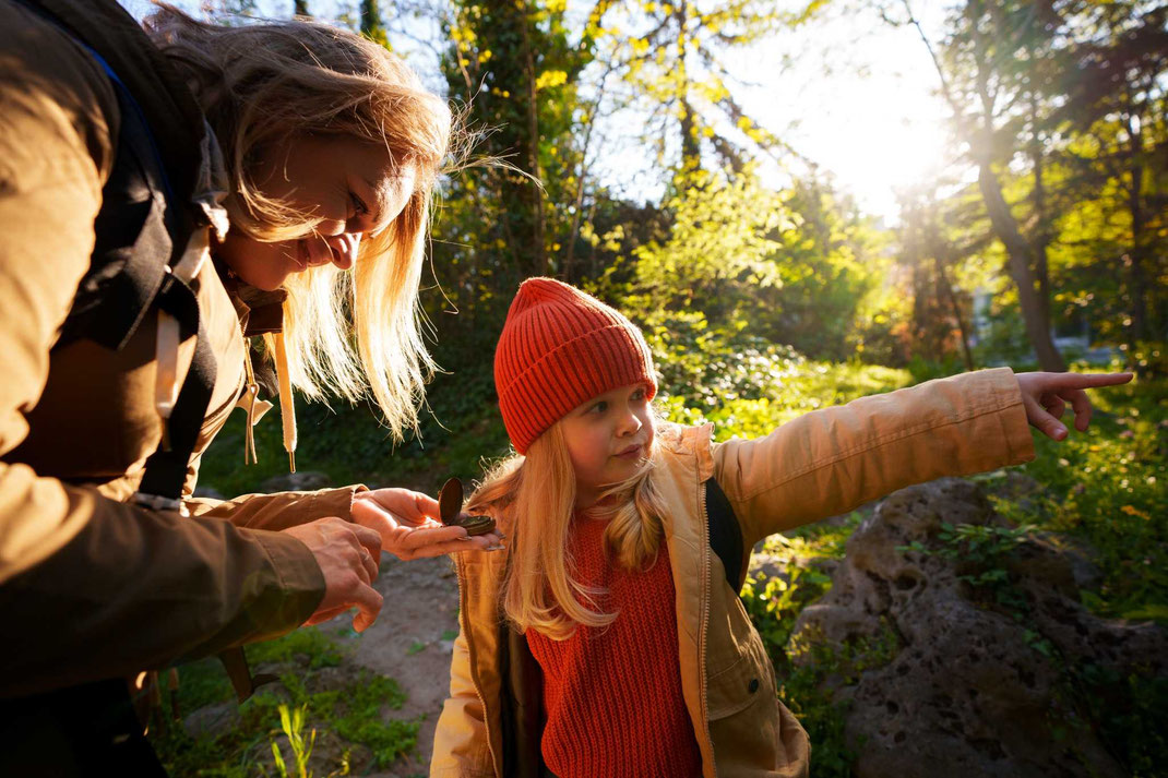 Mutter und Tochter erkunden bei Sonnenlicht den Wald, das Mädchen zeigt neugierig in eine Richtung.