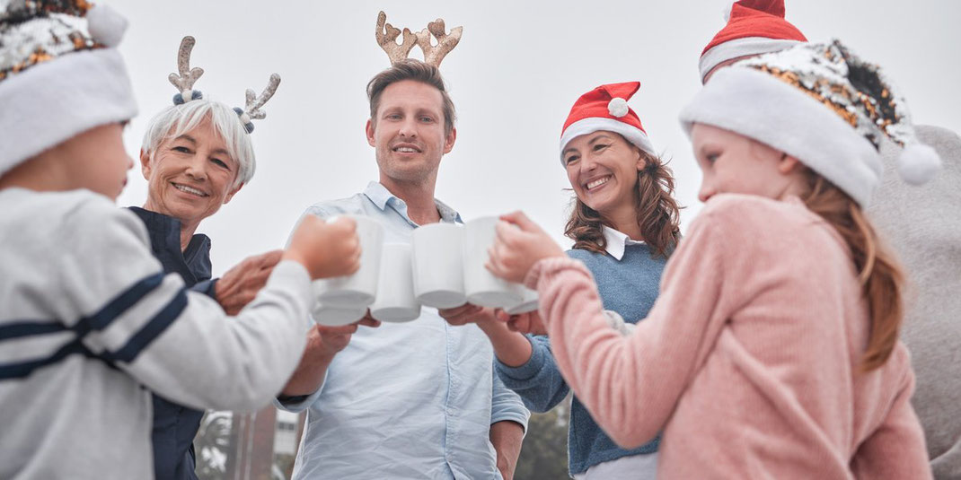 Mehrgenerationen-Familie mit Weihnachtsmützen stößt im Freien mit Tassen an und genießt gemeinsame Zeit