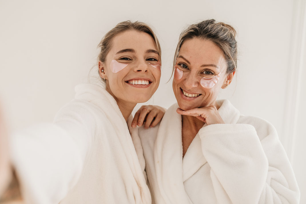 Zwei lächelnde Frauen in weißen Bademänteln mit rosafarbenen Augenpads posieren für ein Selfie – Hautpflege und Freundschaftsmoment im Spa.