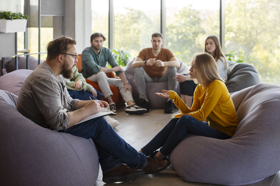 Gruppe von Menschen sitzt in einem hellen Raum auf Sitzsäcken im Gespräch mit einem Therapeuten – offene Atmosphäre bei einer Gruppensitzung oder Selbsthilfegruppe.