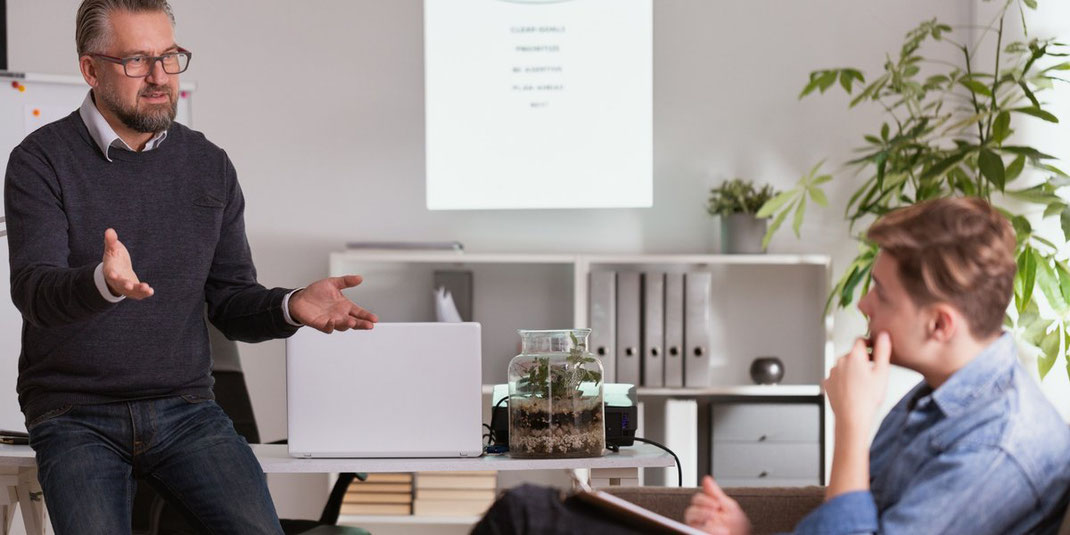 Älterer Mann präsentiert Zeitmanagement-Tipps in einem Büro, junger Zuhörer sitzt nachdenklich mit Notizblock