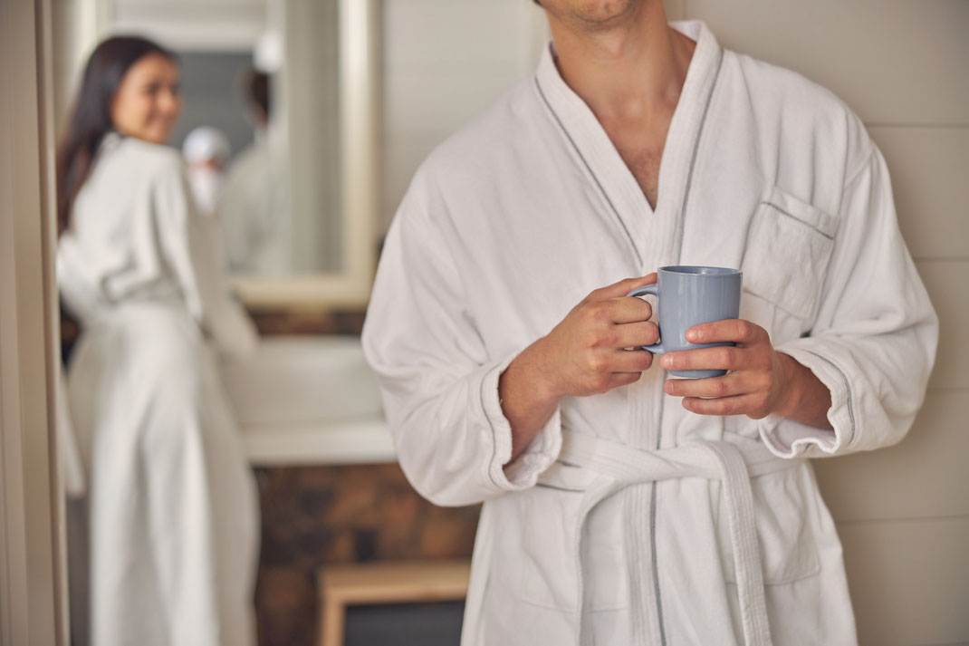 Mann im weißen Bademantel mit Teetasse in der Hand, Frau lächelt im Hintergrund – Morgenroutine, Wellnessgefühl und entspannter Start in den Tag