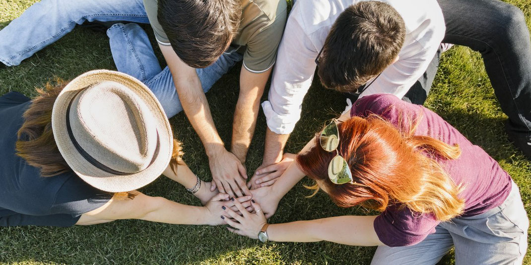 Vier Personen sitzen im Gras und legen ihre Hände in der Mitte zusammen, Ausdruck von Freundschaft und Gemeinschaft.