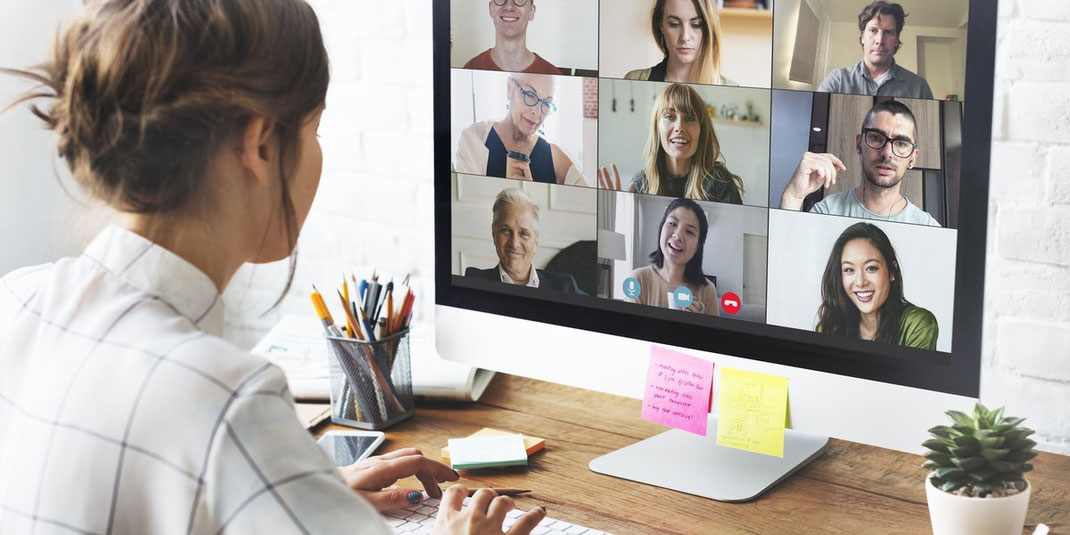 Frau sitzt am Schreibtisch und nimmt an einer Videokonferenz mit mehreren Teilnehmern auf einem großen Monitor teil.