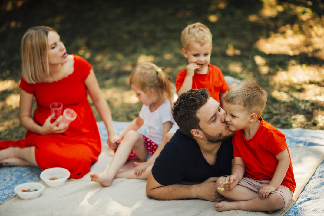 Familie genießt ein gemeinsames Picknick im Park – Vater küsst seinen Sohn, während Mutter und Geschwister entspannt auf der Decke sitzen.
