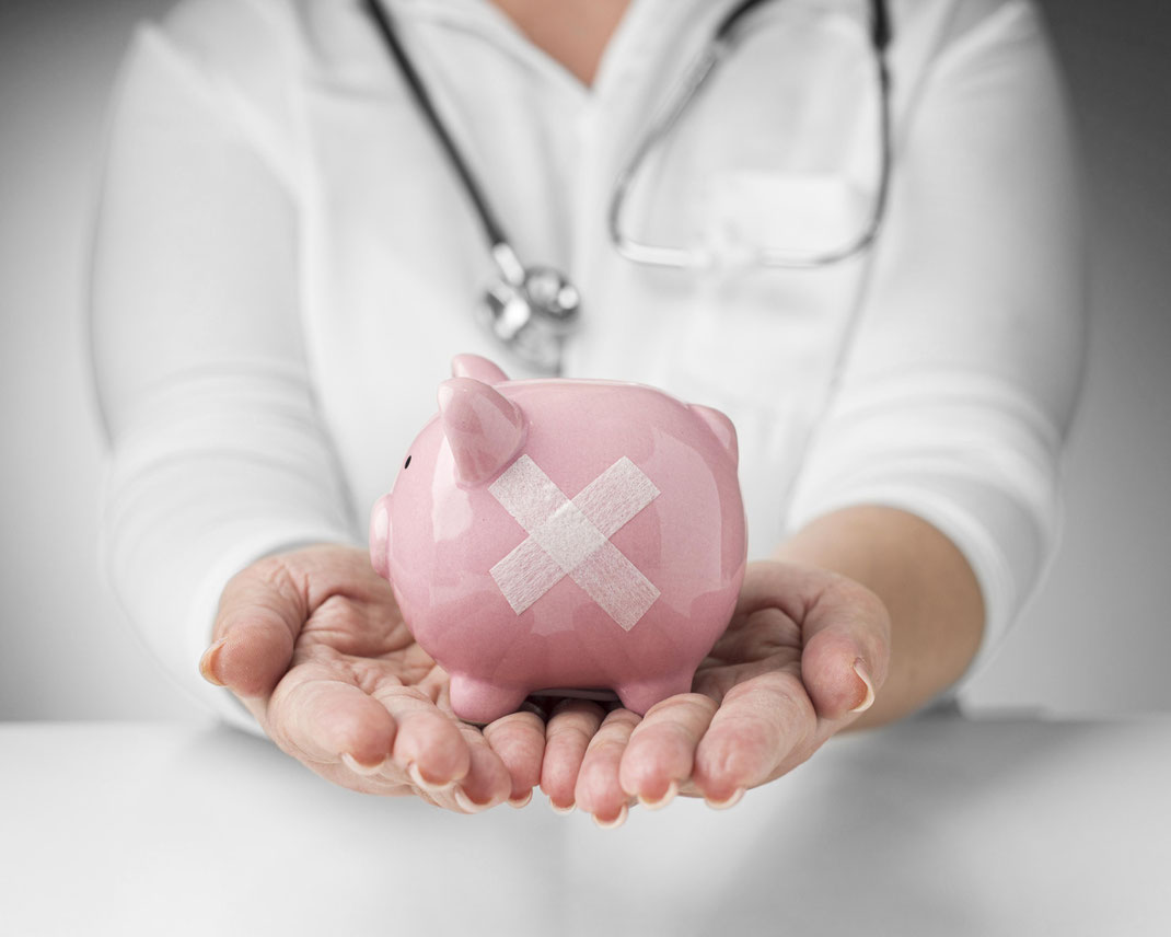 Eine Person in Arztkleidung hält ein rosa Sparschwein mit einem Pflaster – Darstellung der finanziellen Belastung im Gesundheitswesen.
