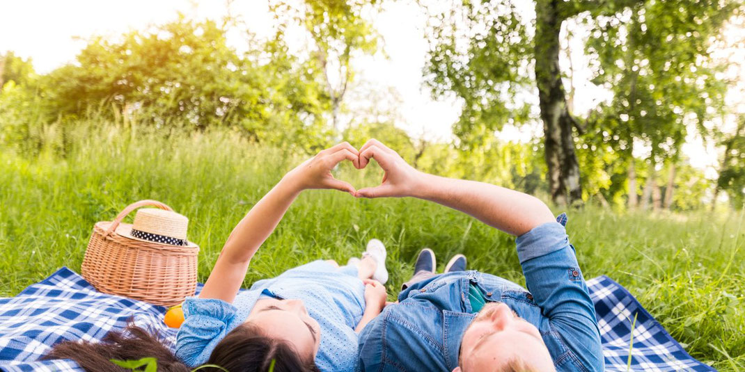 Paar liegt auf einer Picknickdecke im Gras und formt gemeinsam ein Herz mit den Händen