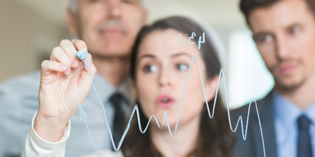 Businessfrau zeichnet ein Diagramm auf eine Glasscheibe, zwei Kollegen im Hintergrund beobachten aufmerksam.