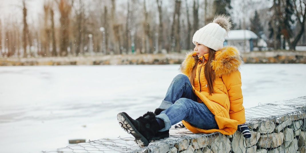 Kind in gelber Winterjacke und weißer Mütze sitzt auf einer Steinmauer im verschneiten Park