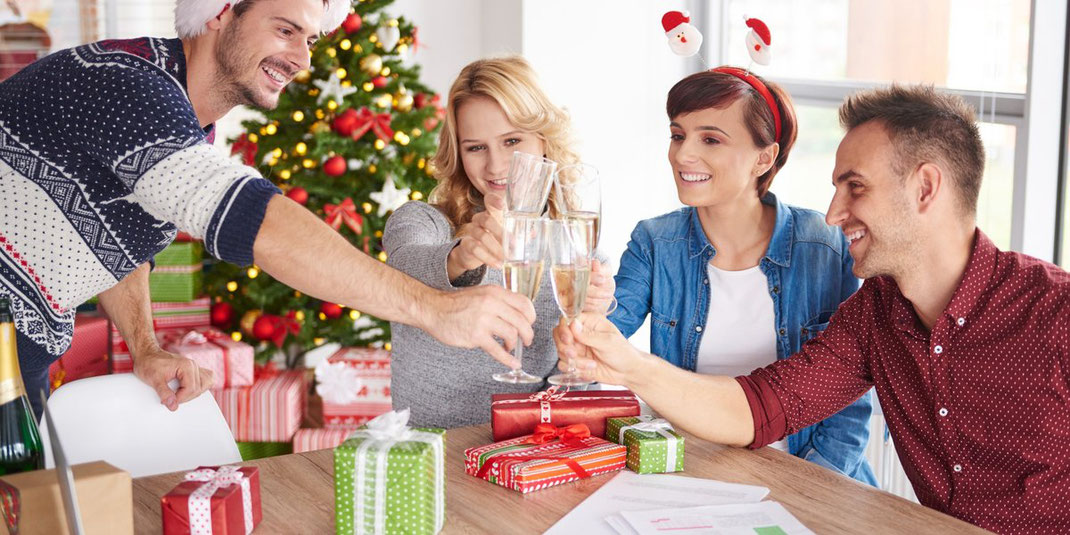 Kolleginnen und Kollegen stoßen bei der Weihnachtsfeier im Büro mit Sekt an, Geschenke und Tannenbaum im Hintergrund