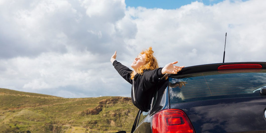 Junge Frau mit offenen Armen lehnt sich aus dem Autofenster und genießt die Landschaft bei bewölktem Himmel