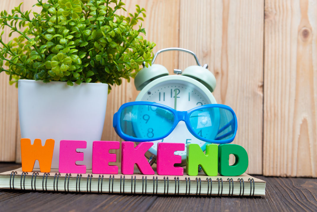 Bunte Buchstaben formen das Wort „WEEKEND“ vor einem Wecker, einer Sonnenbrille und einer Topfpflanze – Darstellung von Vorfreude auf das Wochenende.
