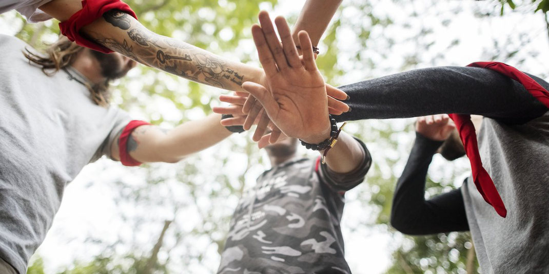 Gruppe von Menschen legt im Kreis ihre Hände übereinander, Symbol für Teamwork und Zusammenhalt im Freien.