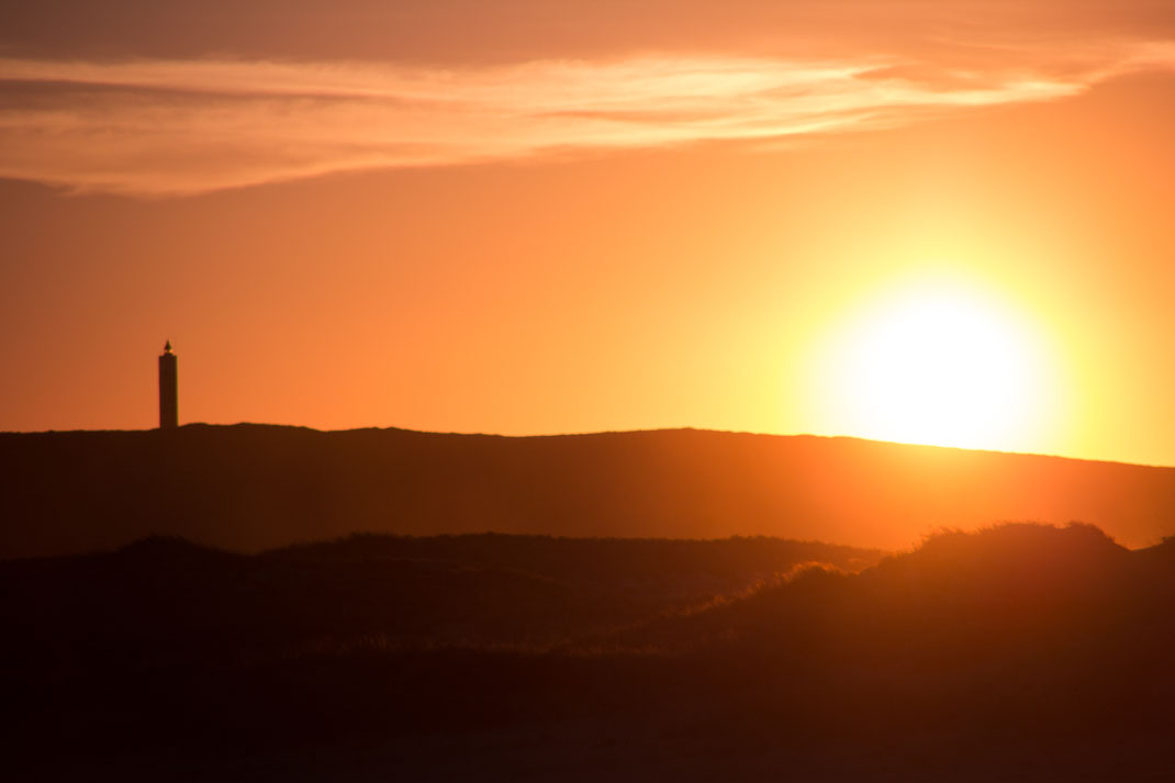 Leuchtturm im Gegenlicht vor einem orangefarbenen Sonnenuntergang über einer Dünenlandschaft