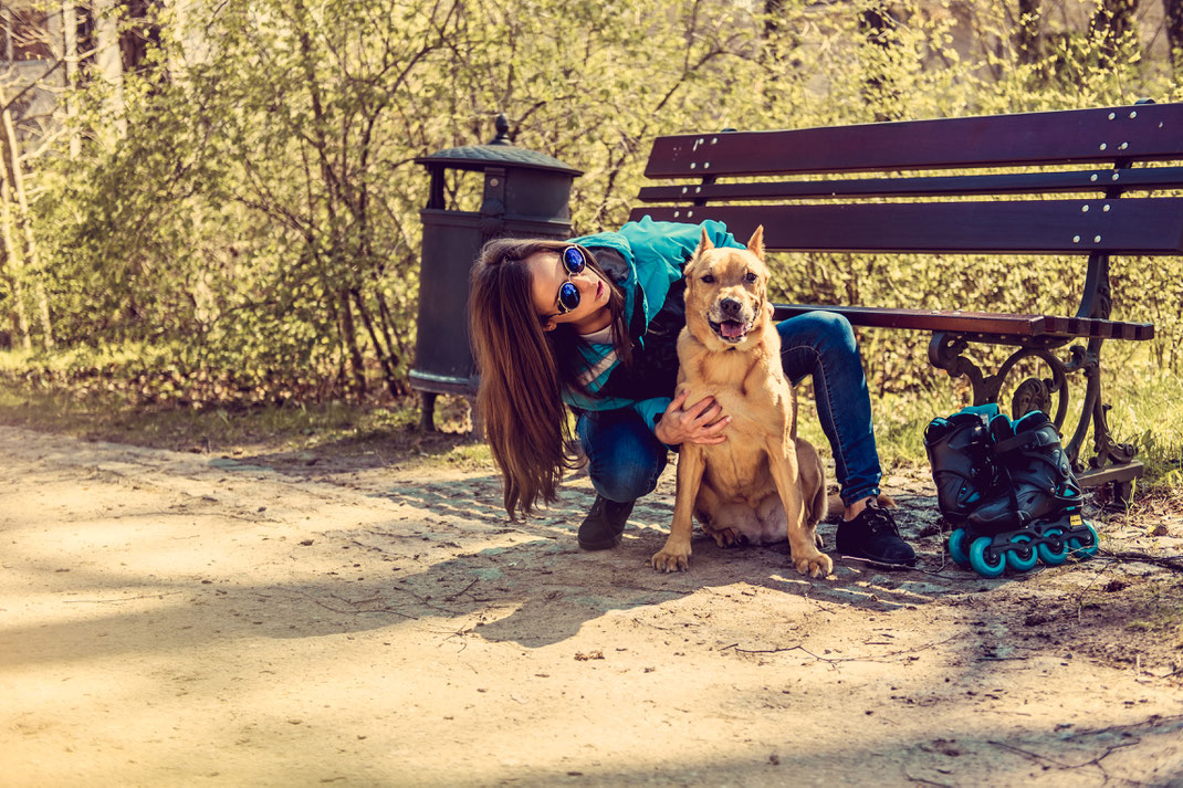 Junge Frau mit Sonnenbrille und blauer Jacke kuschelt fröhlich mit ihrem Hund auf einer Parkbank – neben ihr stehen Inlineskates, im Hintergrund Frühlingsbäume.