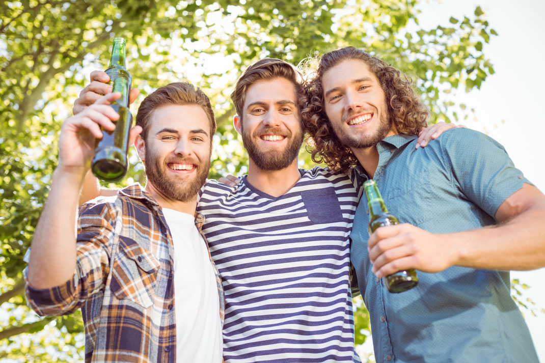 Drei junge Männer mit Bart stehen Arm in Arm im Grünen, halten Bierflaschen in der Hand und lächeln in die Kamera – entspannte Stimmung unter Freunden an einem sonnigen Tag.