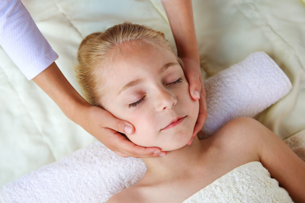 Ein entspanntes Kind mit geschlossenen Augen erhält eine sanfte Gesichtsmassage auf einem Handtuch – Wellnessbehandlung für junge Gäste.