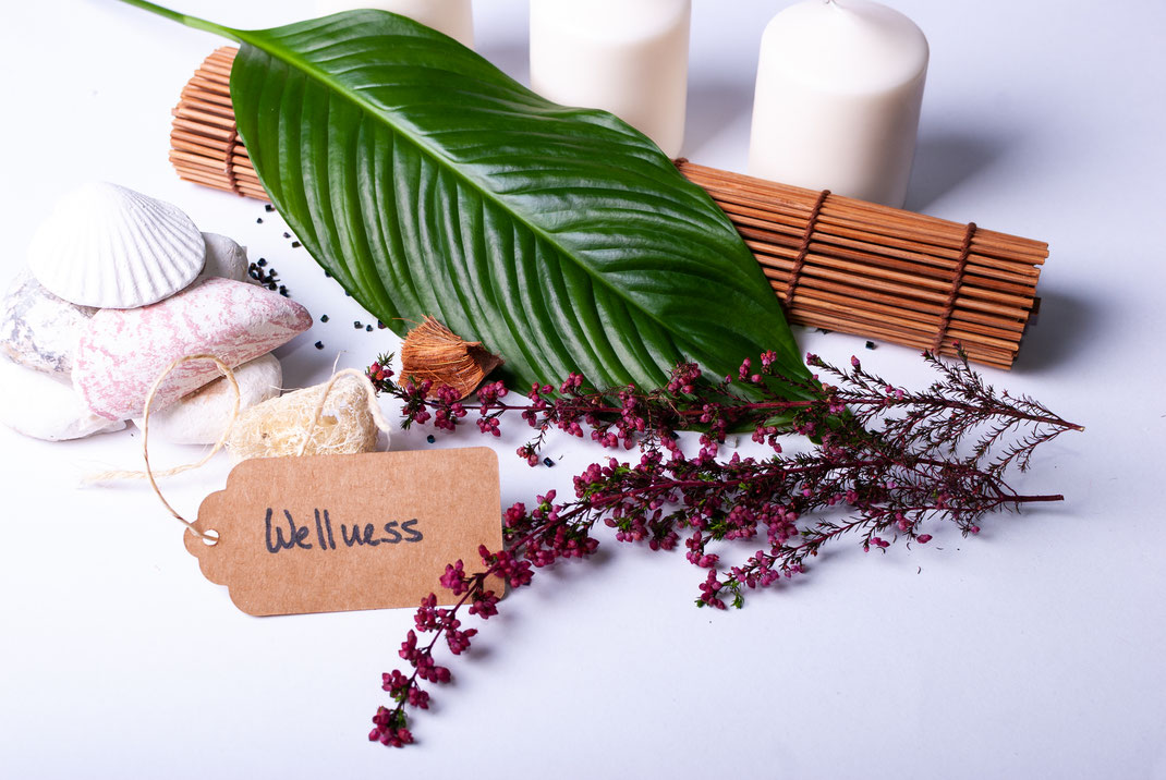 Wellness-Dekoration mit Kerzen, Muscheln, Blüten, Bambusmatte und einem großen grünen Blatt – Symbol für Entspannung und natürliche Pflege.