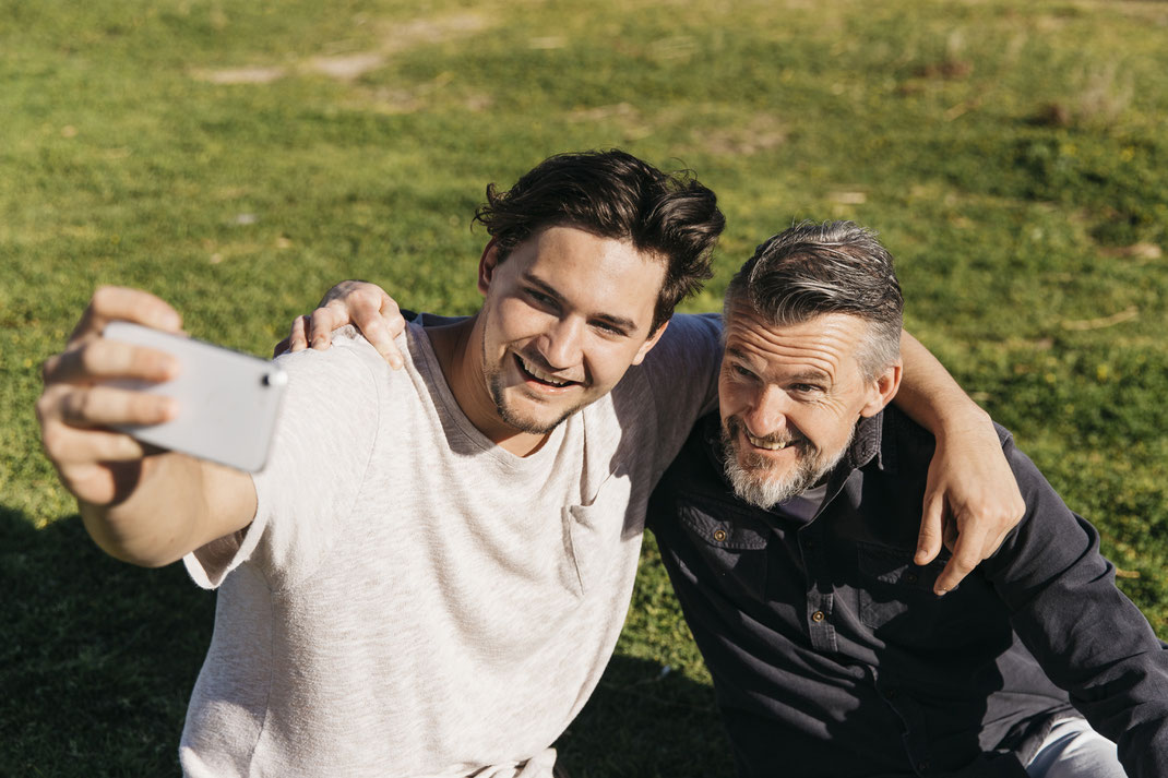 Jüngerer Mann und älterer Mann sitzen Arm in Arm auf einer Wiese und machen gemeinsam ein Selfie – fröhlicher Moment zwischen Vater und Sohn bei schönem Wetter.