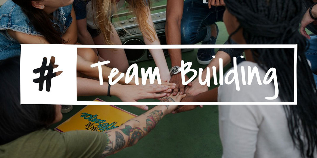 Junge Menschen legen bei einem Teambuilding-Event die Hände zusammen, überlagert von einem Schriftzug mit #TeamBuilding