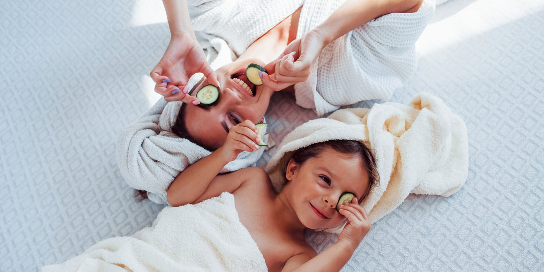 Mutter und Tochter liegen lachend im Bademantel mit Handtuch auf dem Kopf und legen Gurkenscheiben auf die Augen.