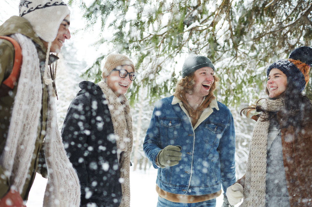 Vier junge Erwachsene lachen gemeinsam unter schneebedeckten Tannen, eingehüllt in warme Winterkleidung – lebendige Szene voller Freundschaft und winterlicher Freude.