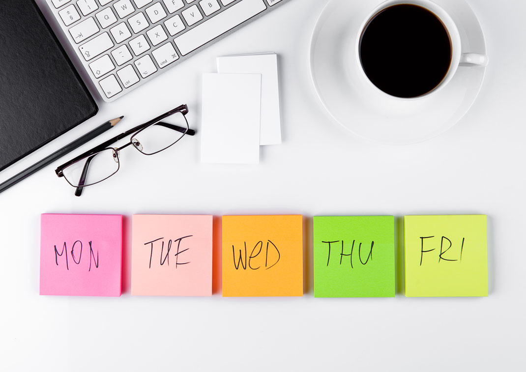 Arbeitsplatz mit Tastatur, Brille, Kaffeetasse und farbigen Notizzetteln für Montag bis Freitag – Symbol für Wochenplanung im Büro.