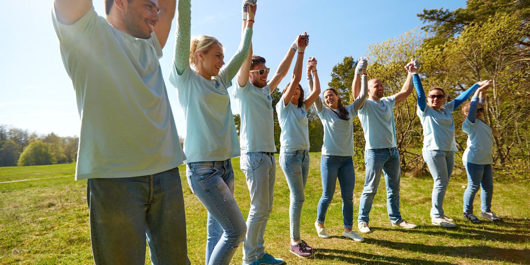 Gruppe von Freiwilligen in blauen Shirts hält sich im Freien an den Händen und hebt sie gemeinsam in die Luft