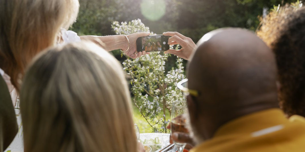 Gruppe von Freunden sitzt draußen am Tisch und macht gemeinsam ein Selfie mit dem Smartphone, sonnige Stimmung im Garten.