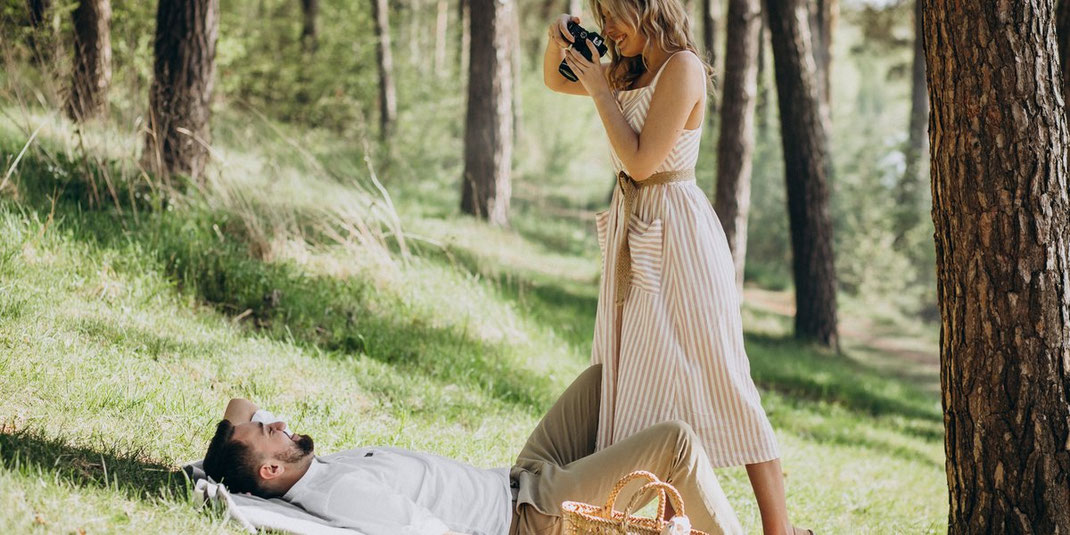 Frau im Sommerkleid fotografiert ihren entspannten Partner beim Picknick im Wald, romantische Stimmung in der Natur.