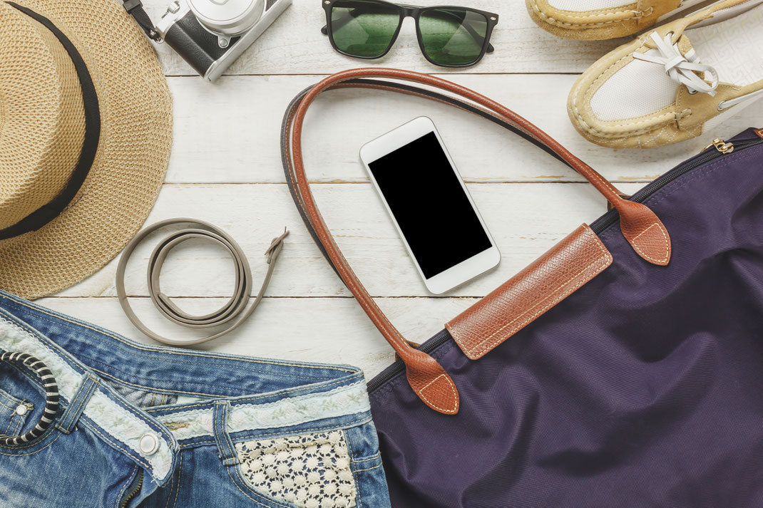 Sommerliche Reiseutensilien wie Strohhut, Sonnenbrille, Smartphone, Handtasche und Jeansshorts liegen auf einem weißen Holztisch – stilvolle Urlaubsvorbereitung.