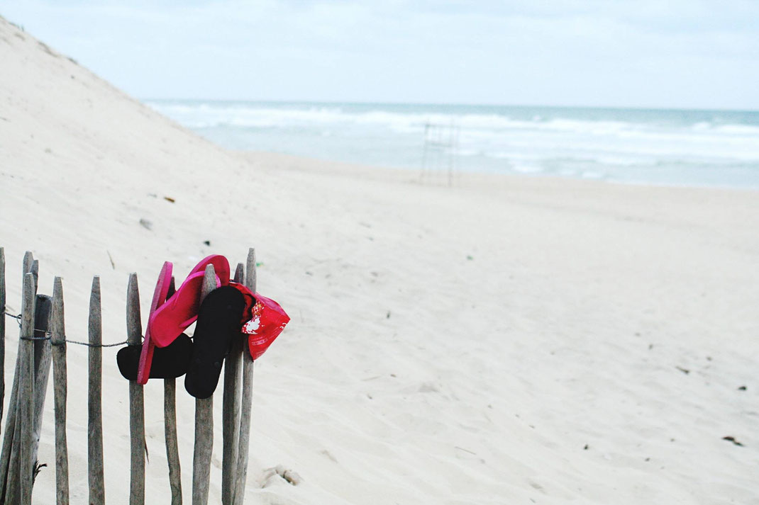 Rote Flip-Flops hängen an einem Holzzaun im Sand mit Blick auf das Meer und sanfte Wellen.