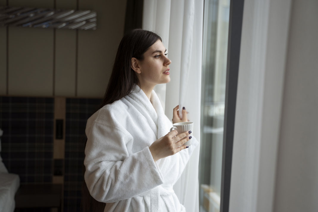 Junge Frau im weißen Bademantel genießt ihren Morgenkaffee am Fenster eines Hotelzimmers – Entspannung, Morgenroutine und Auszeit im Wellnessurlaub