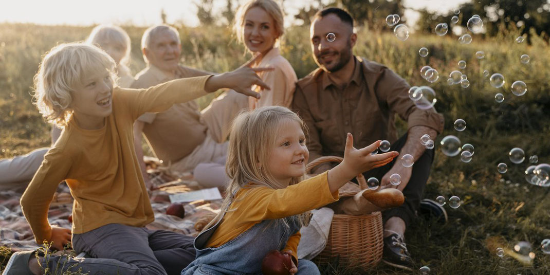 Mehrere Generationen einer Familie sitzen bei einem Picknick auf der Wiese, Kinder spielen fröhlich mit Seifenblasen.