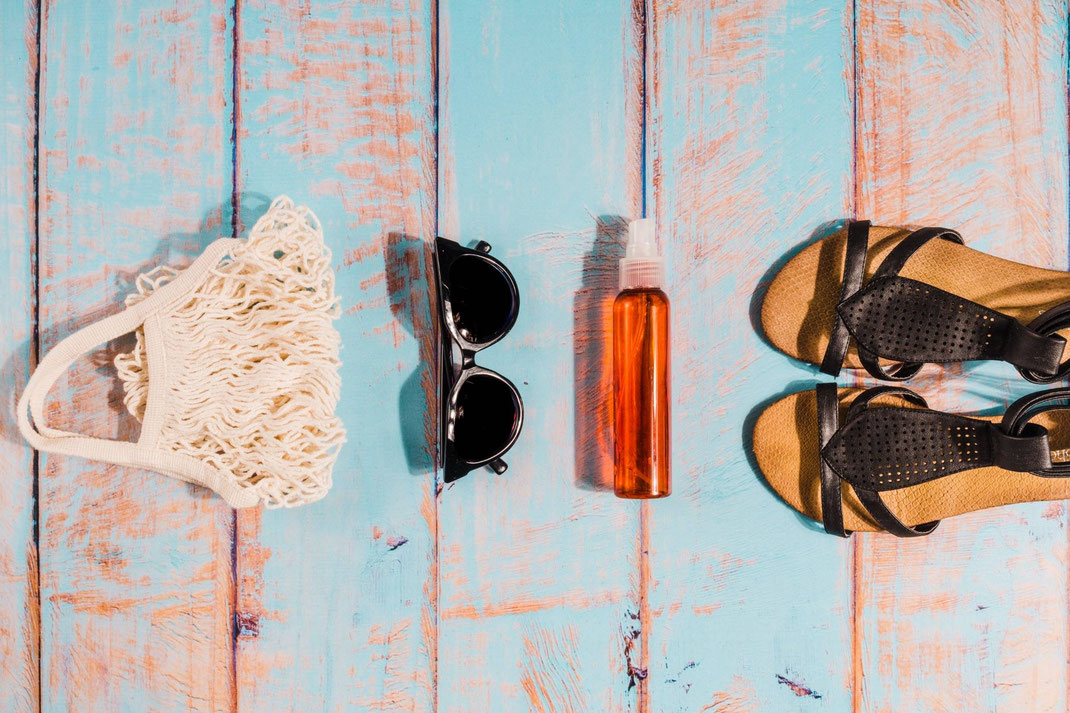 Sommer-Accessoires wie Tasche, Sonnenbrille, Sprayflasche und Sandalen liegen ordentlich auf einem blauen Holztisch.