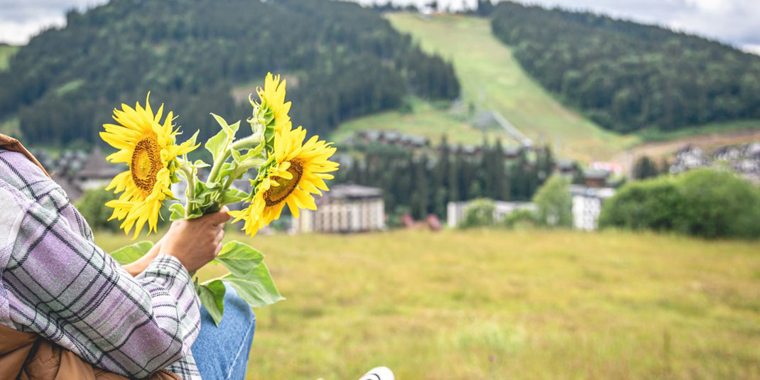 Frau sitzt auf einer Wiese und hält einen Strauß Sonnenblumen mit Berglandschaft im Hintergrund.