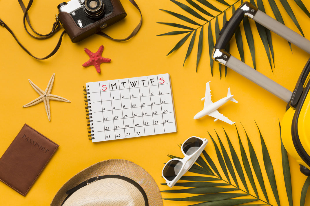Flach gelegte Reiseutensilien auf gelbem Hintergrund: Kalender, Sonnenbrille, Strohhut, Spielzeugflugzeug, Kamera, Koffergriff, Reisepass und Deko mit Strandmotiven – Urlaubsplanung und Vorfreude auf Reisen.