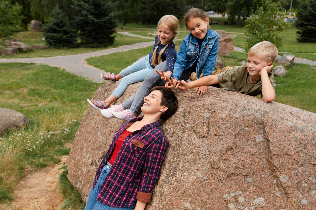 Drei Kinder sitzen auf einem Felsen und spielen fröhlich mit einer Frau, die darunter steht und lacht.