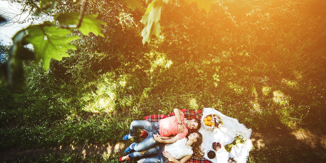Paar liegt entspannt auf einer Picknickdecke im Grünen bei Sonnenlicht.