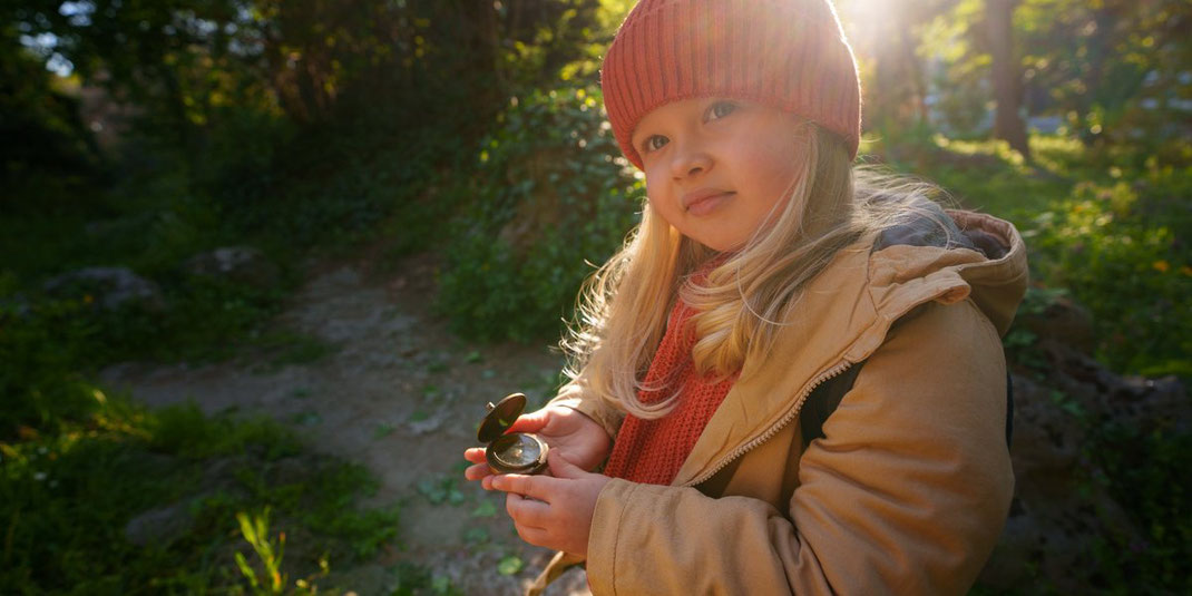 Kleines Mädchen in warmer Kleidung hält einen Kompass beim Erkunden des Waldes im Gegenlicht der Sonne