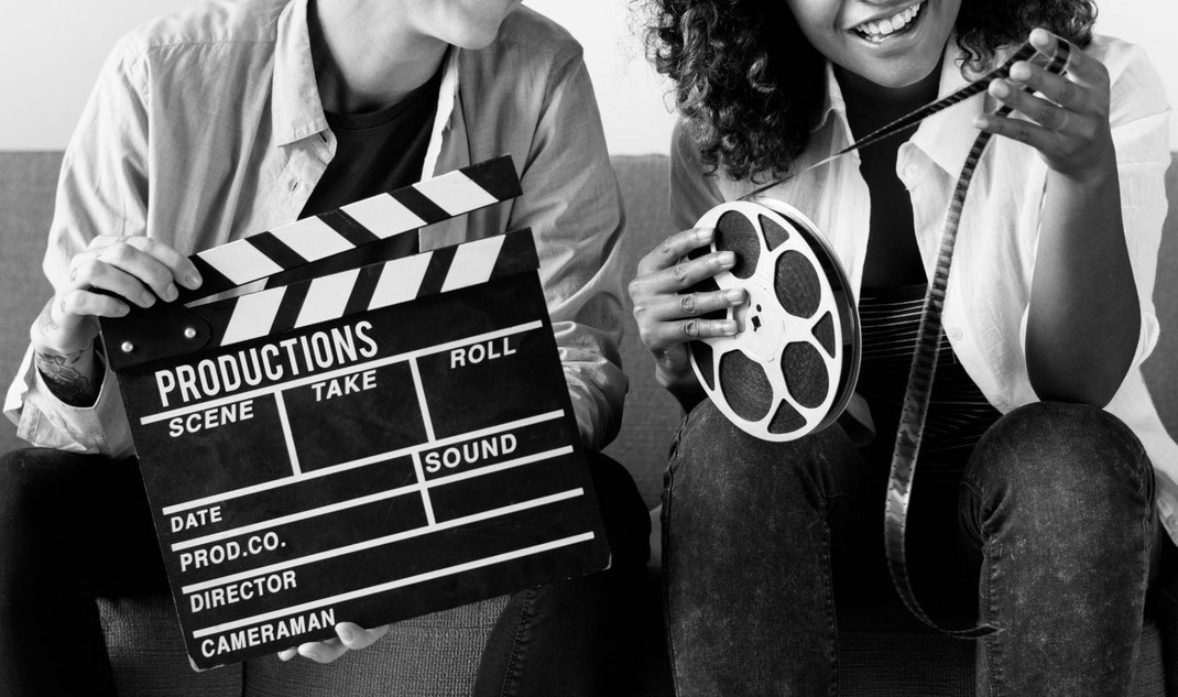 Zwei Personen sitzen auf einem Sofa, halten eine Filmklappe und eine Filmrolle und bereiten sich auf eine Aufnahme vor.