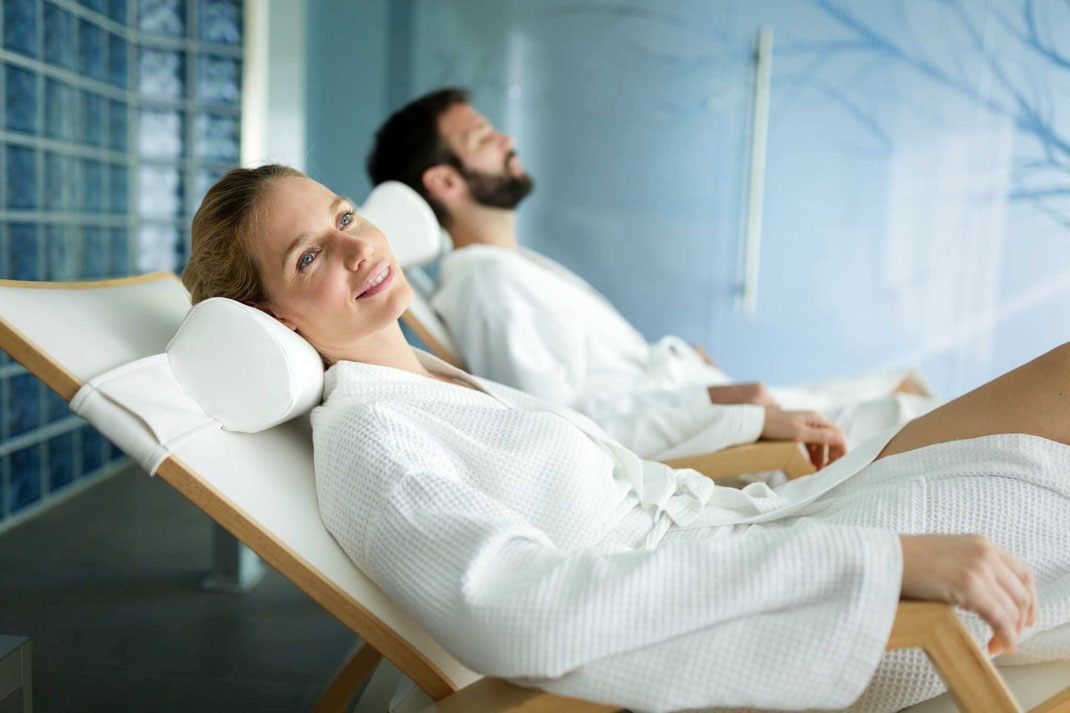 Entspanntes Paar in weißen Bademänteln liegt auf Relaxliegen in einem modernen Spa-Bereich.