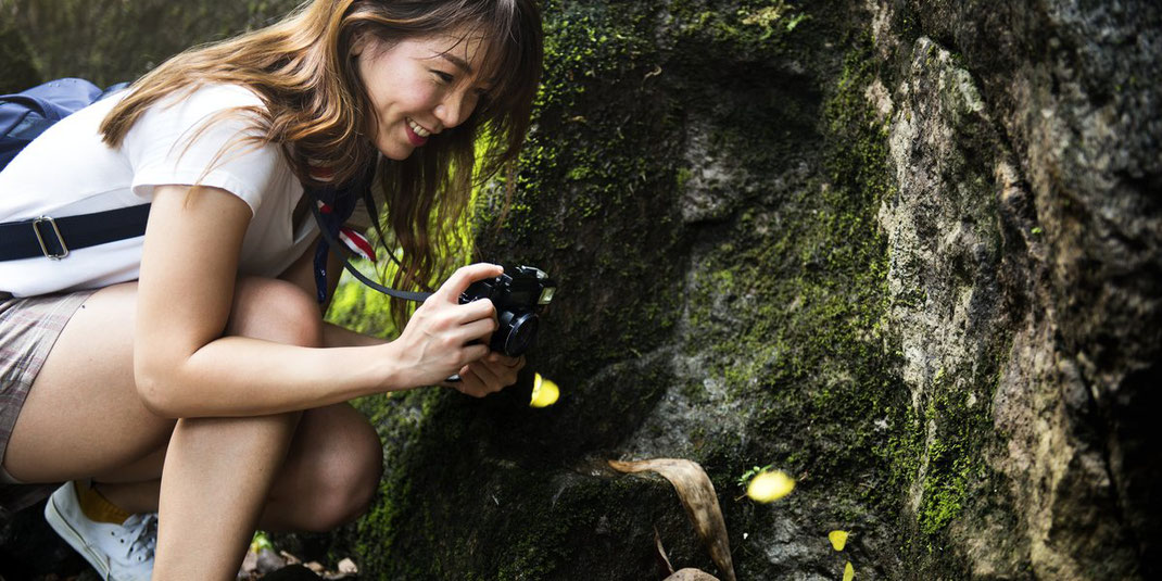 Junge Frau mit Kamera fotografiert nahe einer Felswand im Wald, lächelnd und konzentriert bei der Naturbeobachtung.
