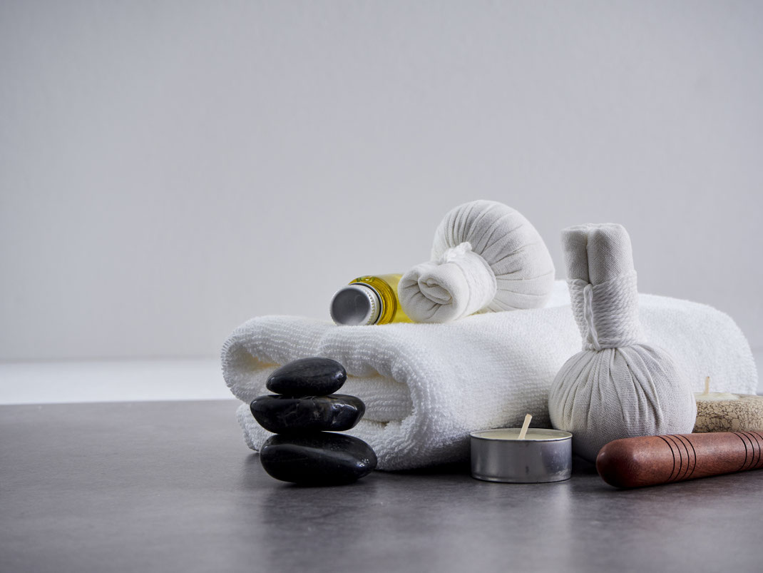 Arrangement mit weißen Handtüchern, Massagekräuterstempeln, schwarzen Steinen, Teelicht und Massageöl – minimalistische Spa-Atmosphäre.